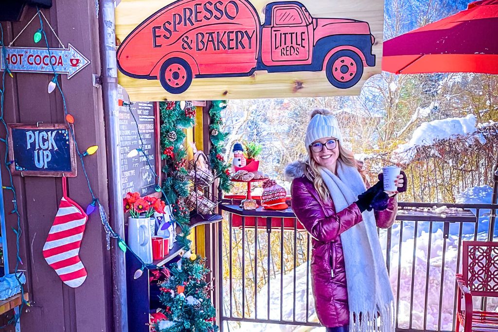 Kate from Kate's Crossing Blog enjoying a warm drink in front of a festive café, "Little Reds Espresso & Bakery." She's bundled up in a purple coat, white scarf, and knit hat, standing next to holiday decorations and a sign for hot cocoa.