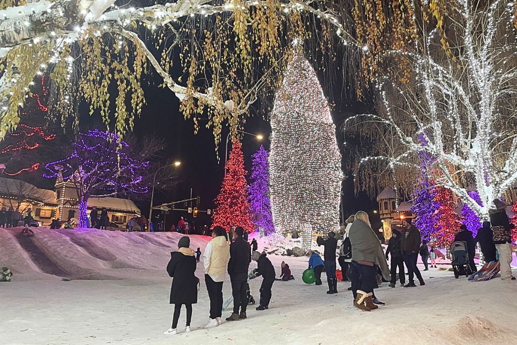  a festive winter evening in Leavenworth, with people gathered around a park filled with vibrant, illuminated trees in red, purple, and white lights. The snow-covered ground and glowing trees create a magical holiday scene. 