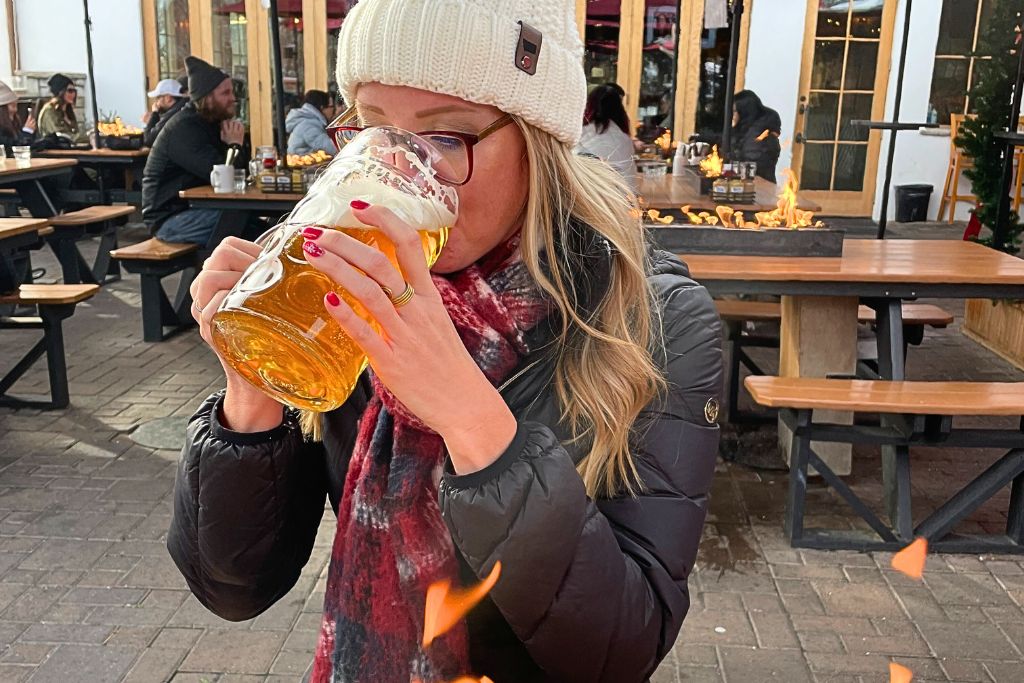 Kate from Kate's Crossing Blog enjoying a large stein of beer at an outdoor seating area, bundled up in a winter coat, scarf, and knit hat. Fire pits and picnic tables surround the area, creating a cozy and warm atmosphere. Savoring a beer at one of Leavenworth's outdoor beer gardens is a relaxing and enjoyable thing to do in Leavenworth in winter, offering a taste of Bavarian culture even in the colder months.