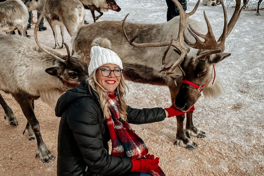 Kate from Kate's Crossing Blog, dressed in a white knit hat, plaid scarf, and red gloves, is petting a reindeer with a red halter in a snowy setting. Surrounded by more reindeer, the scene highlights one of the fun things to do in Leavenworth in winter—visiting reindeer farms for an up-close, festive experience.