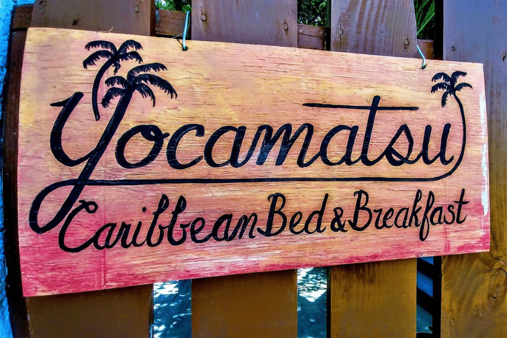 A wooden sign for Yocamatsu Caribbean Bed & Breakfast, featuring a warm gradient background with black lettering and small palm tree illustrations. The sign conveys a tropical, inviting vibe, ideal for a cozy and authentic Caribbean stay in Caye Caulker.