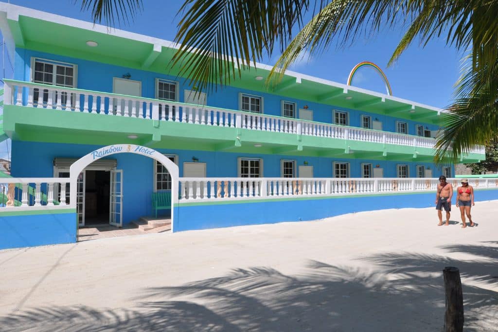 Rainbow Hotel in Caye Caulker stands out with its vibrant blue and green exterior, complete with a rainbow arch above the building, capturing a cheerful, tropical vibe. The two-story structure features wraparound balconies, providing guests with a welcoming seaside ambiance, while palm trees cast shadows across the sandy path in front.