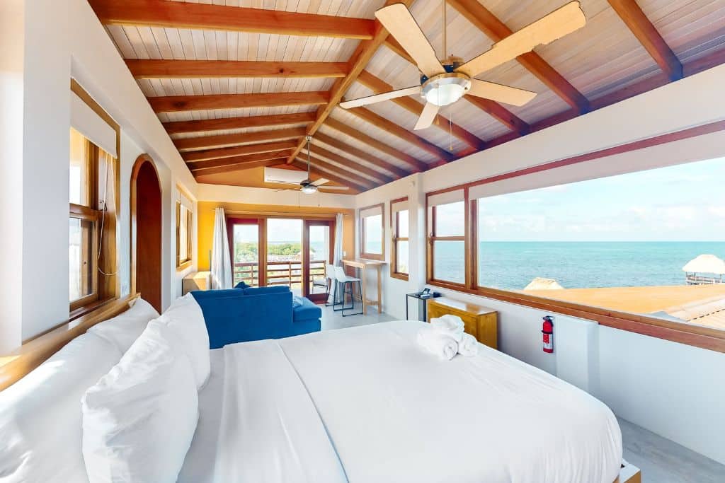 A bright and airy panoramic loft with a scenic balcony overlooking the Caribbean Sea, featuring large windows that fill the room with natural light. The cozy space includes a plush bed, ceiling fans, and a seating area, all framed by a vaulted wooden ceiling, creating a serene and inviting atmosphere for relaxation and breathtaking ocean views.