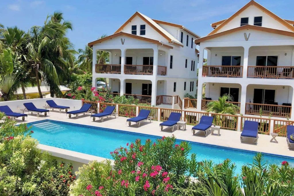 The Velento Oceanfront Penthouse's in Caye Caulker offer a luxurious setting with spacious balconies overlooking a pristine swimming pool surrounded by lush tropical gardens. Rows of blue lounge chairs line the pool area, creating a relaxing, resort-style ambiance for guests to enjoy the sun and scenic views of the Caribbean.