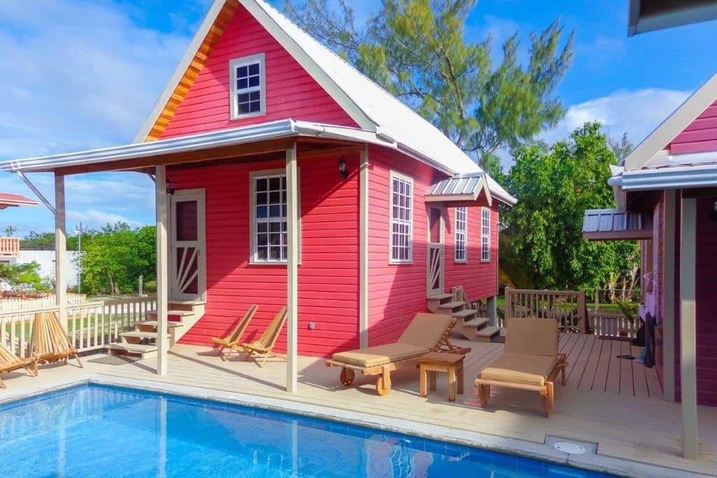 The vibrant Coral Cottage in Caye Caulker features charming pink exteriors and a spacious wooden deck with lounge chairs overlooking a crystal-clear pool. Surrounded by lush greenery, this cozy, colorful retreat offers a relaxed and inviting space for guests to enjoy a tropical getaway.