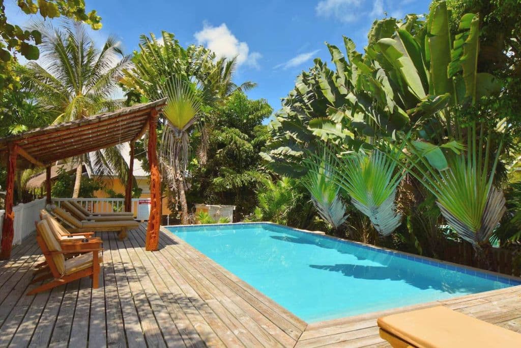 The Hidden Gem Cabana in Caye Caulker offers a secluded poolside retreat surrounded by lush tropical foliage and fan palms. The wooden deck features comfortable lounge chairs under a rustic pergola, creating a tranquil oasis ideal for relaxation and enjoying the natural beauty of the Caribbean.