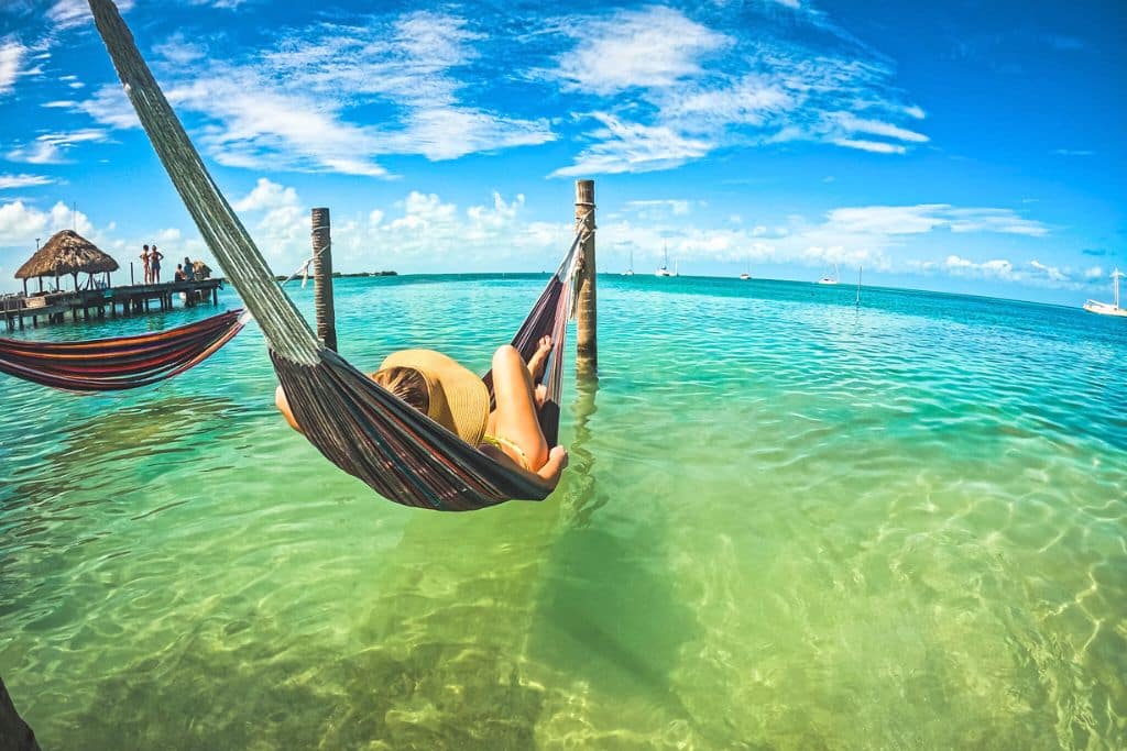 Kate from Kate's Crossing Blog, relaxing in a hammock suspended over the clear turquoise waters of Caye Caulker, enjoying the serene tropical scenery with sailboats in the distance and a bright blue sky above.