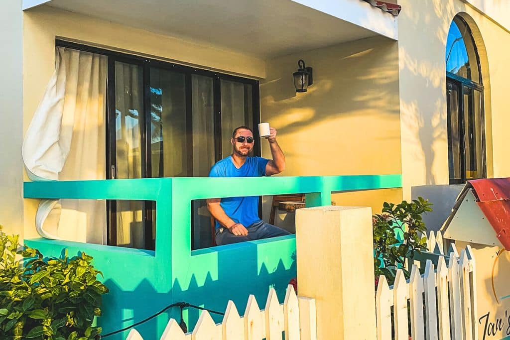 Kate's husband in a blue shirt enjoys his morning coffee on a small balcony in Caye Caulker, surrounded by turquoise accents and framed by a white picket fence. The cozy setting and warm sunlight create a relaxed, welcoming vibe, perfect for a peaceful start to the day in a tropical paradise.