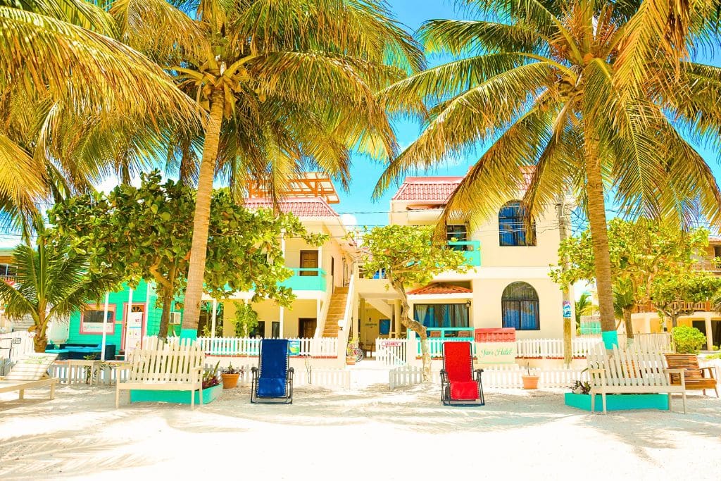 Jan's Hotel in Caye Caulker, Belize, offers a picturesque beachfront view with vibrant turquoise accents, shaded by tall palm trees and surrounded by sandy white pathways. The hotel features comfortable lounge chairs on the beach, creating an inviting space for guests to relax and enjoy the tropical island atmosphere.