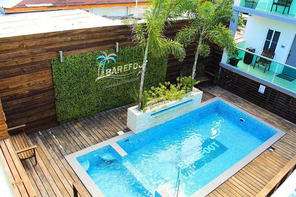 The pool area at Barefoot Caye Caulker Hotel, featuring a modern, blue-tiled pool surrounded by a wooden deck and a lush green wall with the hotel’s logo. Palm trees and seating add to the tropical vibe, offering a private and stylish space for guests to relax and enjoy their stay in Belize.