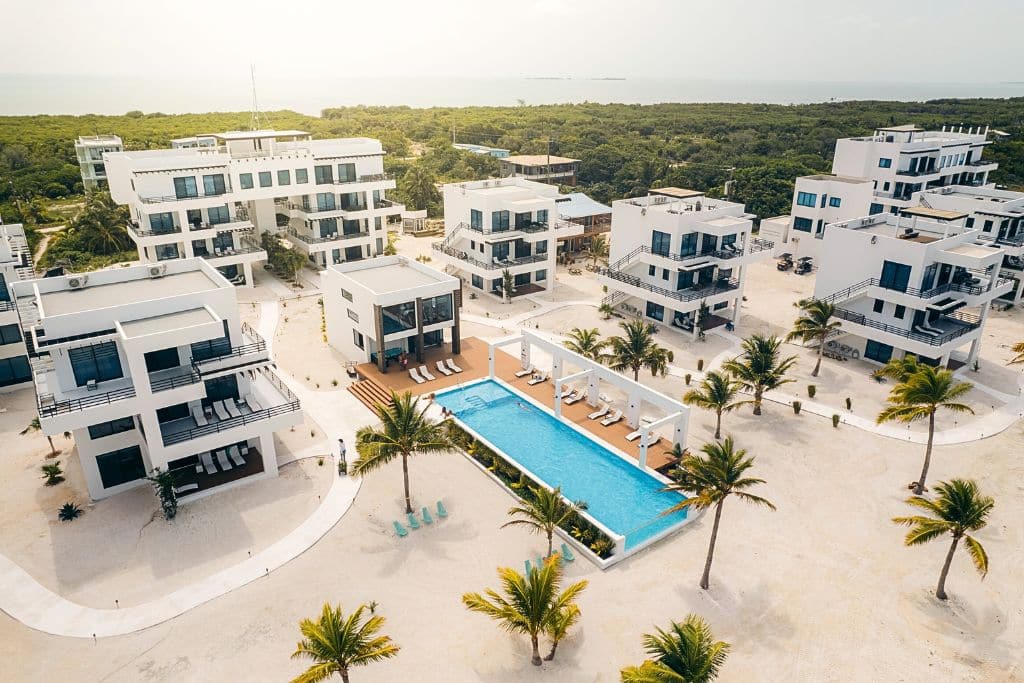 Aerial view of BluZen, a modern luxury resort in Caye Caulker featuring sleek, white multi-story buildings surrounding a long, central swimming pool. The resort’s minimalist design is complemented by palm trees and spacious lounging areas, providing a serene, upscale atmosphere with views of lush greenery and the ocean in the distance.