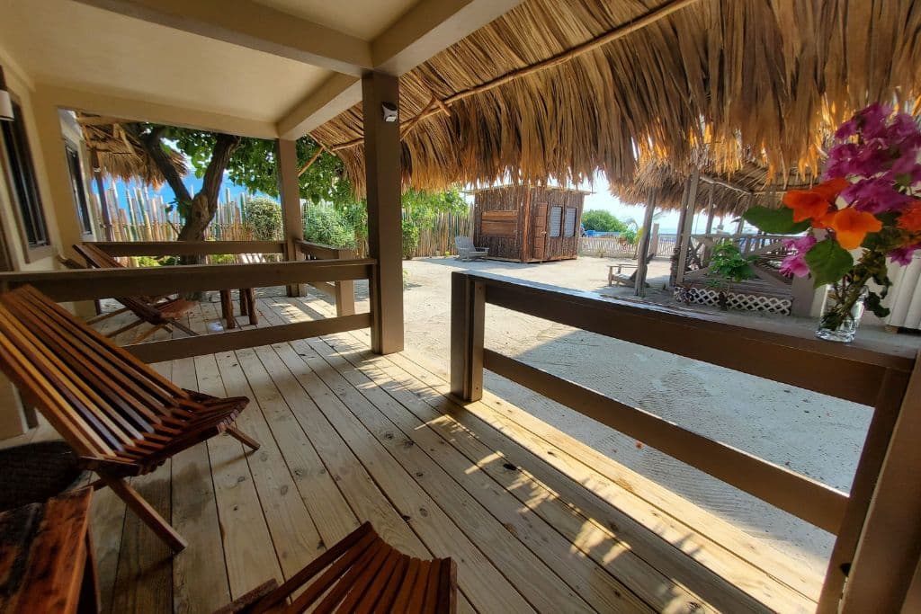 The cozy porch at The Bounty Boutique Bohemian Hotel in Caye Caulker, featuring rustic wooden chairs and a thatched roof that creates a shaded, tropical ambiance. Vibrant flowers in a vase add a splash of color to the natural wood decor, with views of sandy pathways and additional huts in the background, enhancing the relaxed, bohemian vibe.