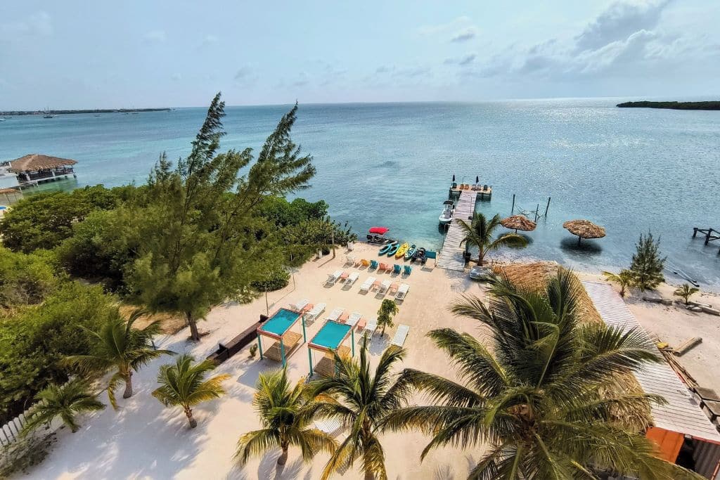 The beachfront view at Bay Towers Hotel in Caye Caulker, showcasing white sand, turquoise cabanas, and lounge chairs overlooking the Caribbean Sea. A wooden pier extends into the water with kayaks, shaded seating, and thatched umbrellas, creating an inviting tropical paradise for guests to enjoy water activities and relaxation.