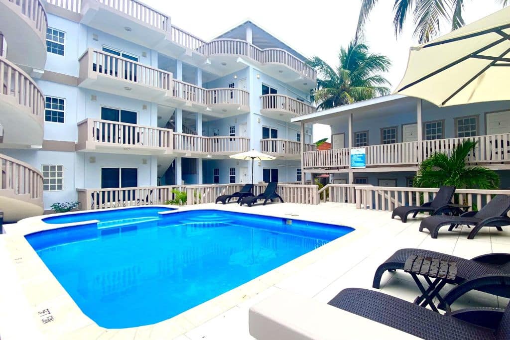 The pool area at La Isla Resort in Caye Caulker, featuring a bright blue swimming pool surrounded by lounge chairs and shaded seating. The multi-story resort buildings with wraparound balconies overlook the pool, offering a comfortable and inviting atmosphere for relaxation in a tropical island setting.