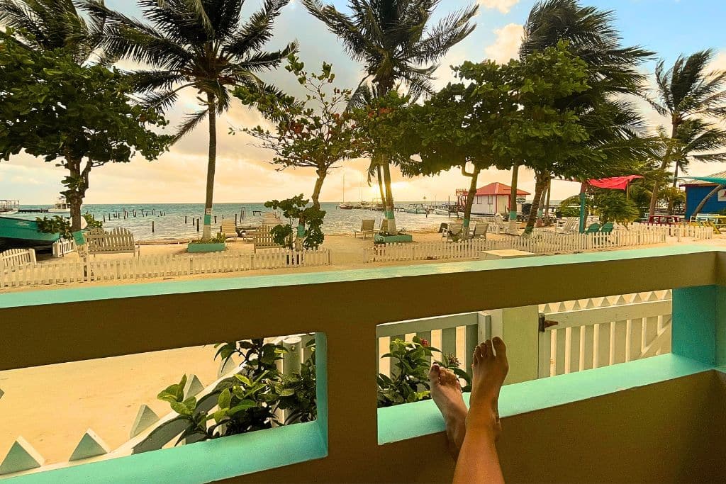 A relaxing view from a porch in Caye Caulker, Belize, looking out at a beach scene with palm trees swaying in the breeze, calm waters, and small piers extending into the ocean. In the foreground, a person’s legs are stretched out, capturing a peaceful moment at a seaside retreat.