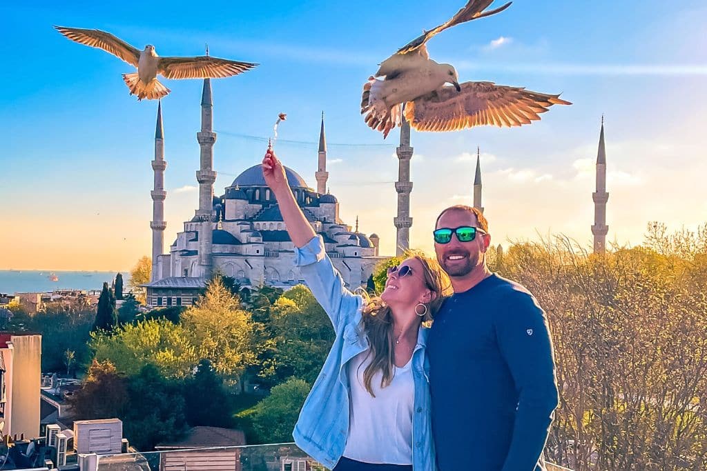 Kate of Kates Crossing Blog and her partner are enjoying a rooftop experience in Istanbul, Turkey, with the iconic Blue Mosque (Sultan Ahmed Mosque) in the background during a sunny day. Kate feeds seagulls mid-flight, capturing a vibrant and lively moment against the backdrop of the mosque's elegant minarets and lush greenery.