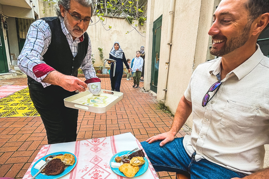 This image shows a local Turkish man offering traditional Turkish tea to a smiling guest seated at an outdoor table. The table is adorned with plates of Turkish sweets and pastries, while the background reveals a cozy courtyard with colorful rugs and other locals enjoying the space. The scene captures a warm and authentic cultural experience in Turkey.