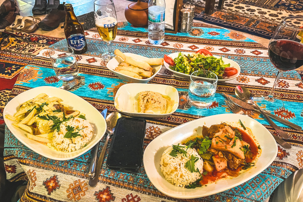 A colorful Turkish meal spread on a patterned tablecloth, featuring dishes like rice, roasted meats, and vegetables alongside fresh salad and bread. Drinks, including a glass of red wine and Efes beer, accompany the feast, with decorative carpets visible in the background.