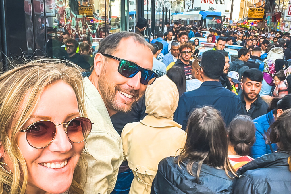 A vibrant street scene in Istanbul Turkey at the end of Ramedan, with a bustling crowd of locals and tourists filling the area. A smiling couple in the foreground enjoys the lively atmosphere, surrounded by a mix of colorful signs and storefronts.