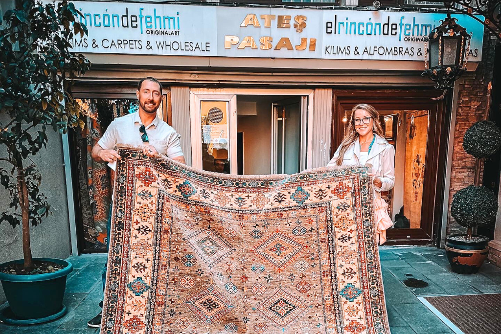 A smiling couple standing in front of a Turkish carpet shop, proudly holding up a beautifully patterned rug. 