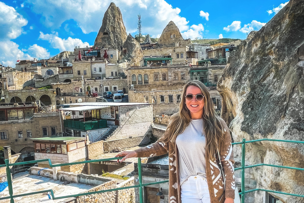 Kate standing on a terrace overlooking the captivating cave dwellings and stone buildings of Göreme, Cappadocia, Turkey. The iconic fairy chimneys rise in the background under a bright blue sky, showcasing the region's unique geological and architectural wonders.