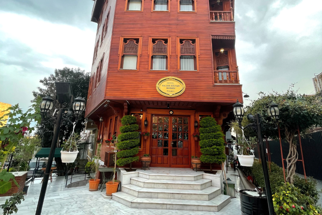 This image showcases a beautifully crafted wooden boutique hotel, adorned with traditional Turkish architectural elements. The charming entrance features neatly trimmed topiary, marble steps, and decorative hanging lanterns, creating an inviting and elegant ambiance.