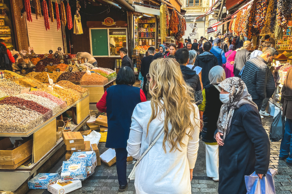 This image captures the lively atmosphere of a bustling Turkish market, with colorful stalls displaying dried fruits, nuts, and spices. Shoppers, including locals and tourists, move through the vibrant street lined with hanging dried peppers and aromatic goods.