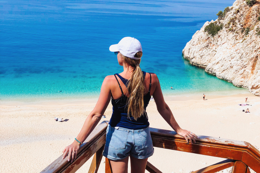 Kate standing on a wooden balcony overlooking the crystal-clear turquoise waters and golden sands of Kaputaş Beach in Turkey. Dressed in casual summer attire with a white cap, she admires the breathtaking coastal scenery.