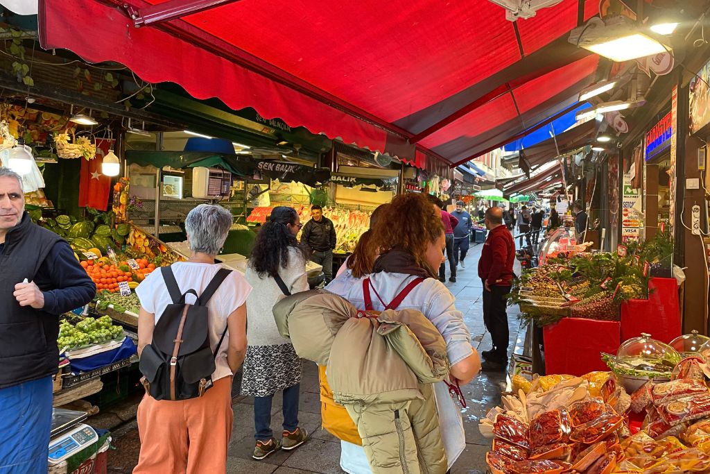 This image captures the bustling atmosphere of a vibrant Turkish market, with colorful stalls displaying fresh produce, spices, and other goods. Shoppers browse under bright red awnings, adding energy and warmth to the scene.