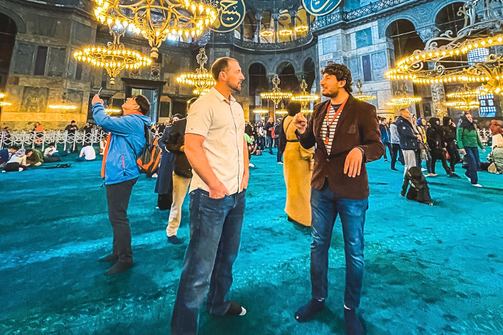 This image captures the interior of the Hagia Sophia mosque in Turkey, with visitors walking or sitting on the richly colored teal carpet under ornate golden chandeliers. A man and a tour guide engage in a conversation in the foreground, while others admire the intricate architecture and take photos. The atmosphere reflects the grandeur and cultural significance of the space.