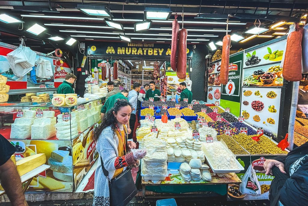 This image showcases a lively Turkish market stall specializing in cheeses, olives, and cured meats, with neatly arranged displays and vibrant colors. Customers browse the selection under bright lighting, while staff assist in the background. The variety of local delicacies highlights the rich culinary culture of Turkey.