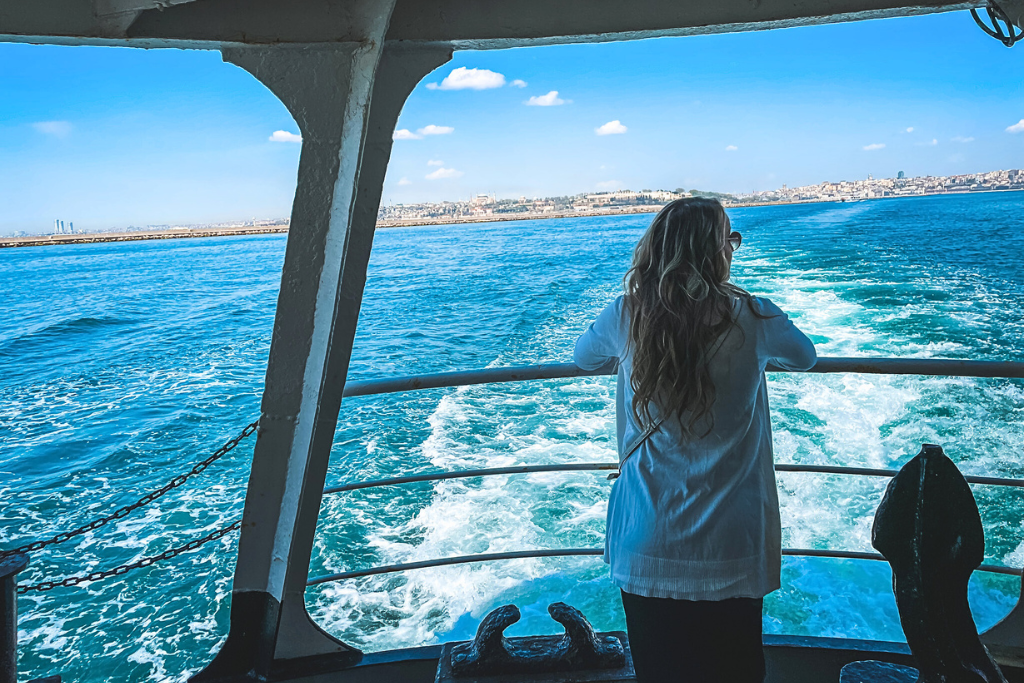 Kate standing at the railing of a ferry, gazing out over the bright blue waters with the wake trailing behind the boat. The distant skyline of a Turkish city is visible under a clear sky, adding a sense of adventure and serenity to the scene.