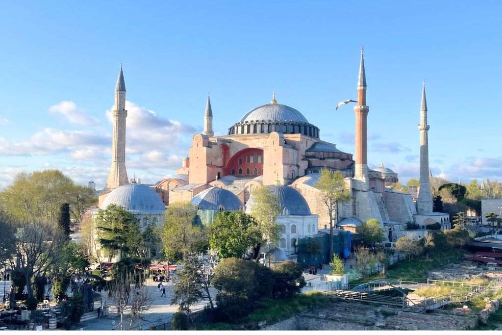 This image captures the iconic Hagia Sophia in Istanbul, Turkey, bathed in soft sunlight under a blue sky. The architectural masterpiece, with its grand dome and towering minarets, stands amidst lush greenery and bustling visitors below.