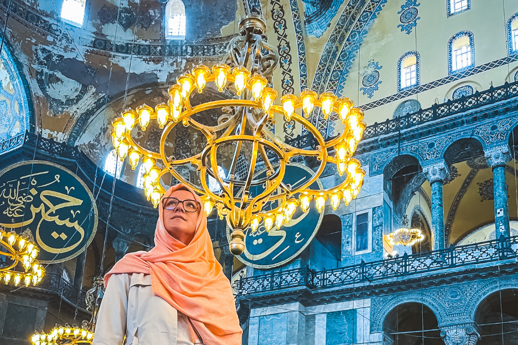 This image features Kate wearing a peach colored headscarf and glasses, standing beneath a grand chandelier inside the Hagia Sophia mosque in Turkey. The intricate architectural details, calligraphy, and vibrant decor of the ceiling and walls create a stunning backdrop.