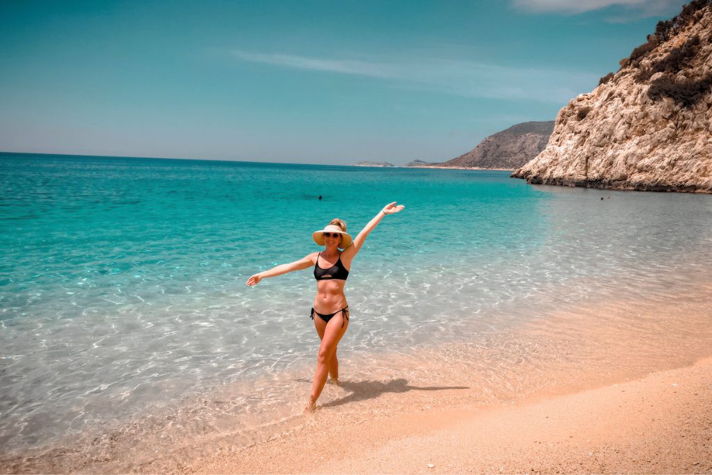 Kate in a black bikini and sun hat enjoying the pristine beach at Kaputaş in Turkey. The crystal-clear turquoise waters and golden sand create a picturesque coastal setting, with rocky cliffs framing the serene scene.