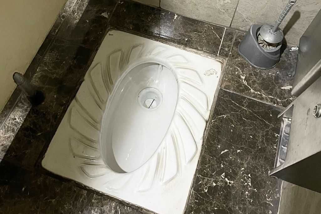 This image features a traditional squat toilet, commonly found in public restrooms in Turkey and other parts of the world. The ceramic fixture is set into a dark marble floor, with a water nozzle and cleaning brush nearby.