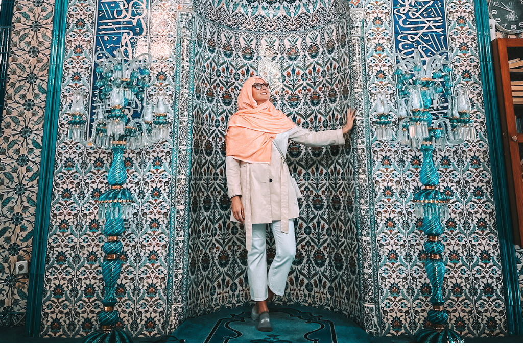 Kate wearing a peach colored headscarf and white clothing standing against an intricately tiled wall inside a mosque in Turkey. The vibrant blue and turquoise patterns, adorned with floral motifs and Arabic calligraphy, create a breathtaking backdrop.