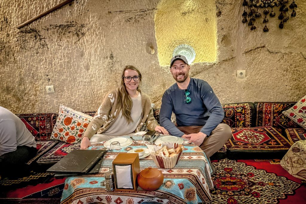 Kate and her partner seated in a cozy, traditional Turkish restaurant, carved into the rocky interior of Cappadocia. They are surrounded by vibrant patterned cushions and rugs, with a table adorned with Turkish-style dishes and decor.