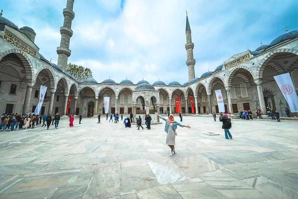 Kate stands in the courtyard of the Blue Mosque (Sultan Ahmed Mosque) in Istanbul, Turkey with her arms stretched out to the side showing of the grandeur. The expansive space is framed by grand arches and towering minarets under a partly cloudy sky, with visitors exploring the historic site.