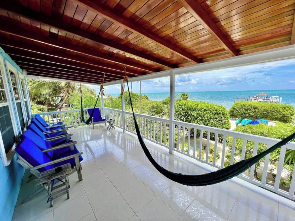 The spacious balcony at Laura's Lookout Oceanfront in Caye Caulker, featuring comfortable blue lounge chairs and hammocks overlooking lush greenery and the Caribbean Sea. The polished tile floor and wooden ceiling create a refined, relaxing ambiance, ideal for enjoying panoramic ocean views and a gentle sea breeze.