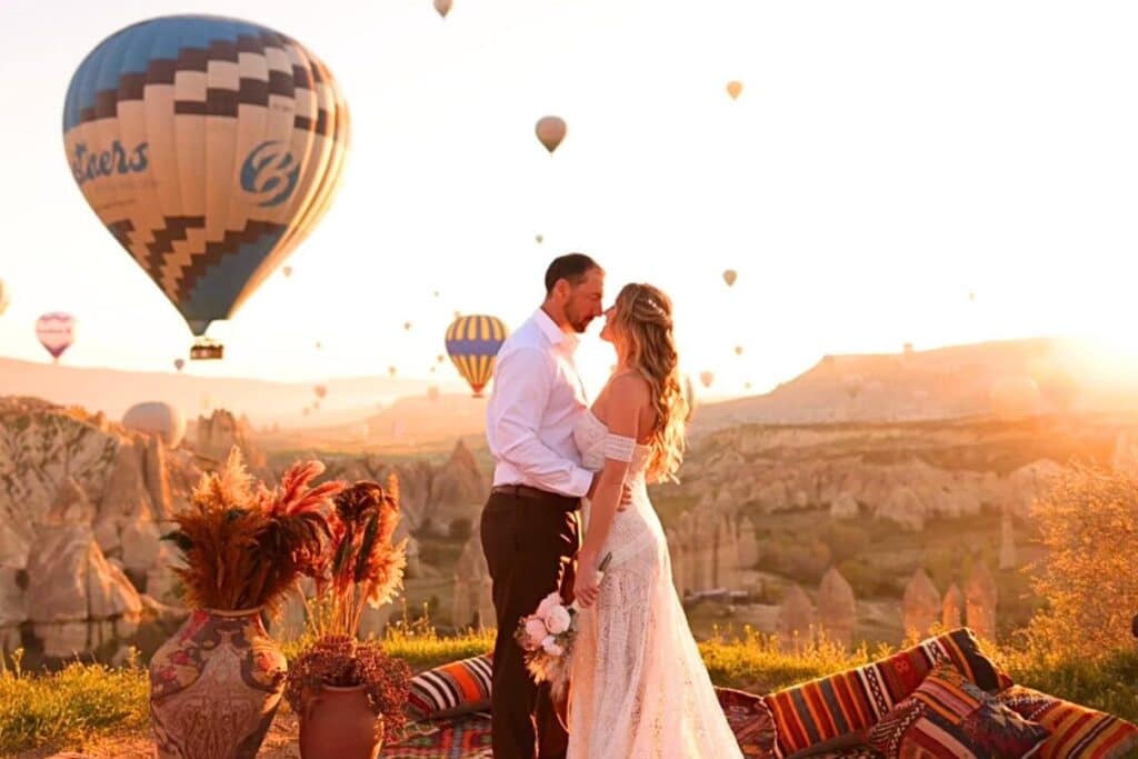 A couple stands on a Turkish rug in Cappadocia at sunrise, sharing an intimate moment surrounded by pottery and dried flowers. The bride wears a white lace dress, and the groom is in a white shirt, with colorful hot air balloons filling the golden-lit sky in the background. The scene captures the region's romantic and scenic charm.