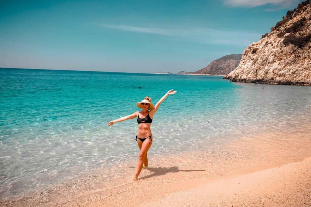 Kate, in a black bikini and wide-brimmed hat, poses playfully on a sandy beach along the turquoise waters of the Turkish Riviera. Rugged cliffs and a calm sea extend into the background under a clear blue sky.