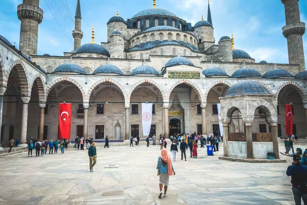 Visitors walk through the grand courtyard of the Blue Mosque (Sultan Ahmed Mosque) in Istanbul, with its iconic domes, arches, and minarets under a cloudy sky. The courtyard features Turkish flags and intricate architectural details. Kate in a light pink headscarf and denim jacket is prominently seen walking toward the entrance.