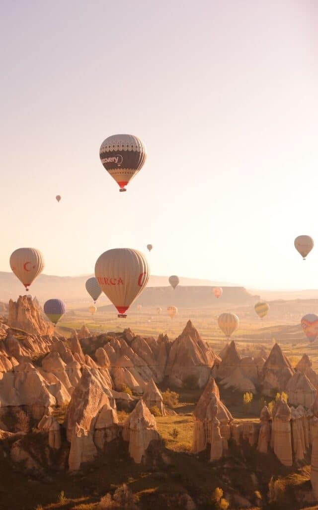 Hot air balloons float gracefully over the unique rock formations and fairy chimneys of Cappadocia at sunrise. The warm golden light illuminates the landscape, enhancing the earthy tones of the terrain.