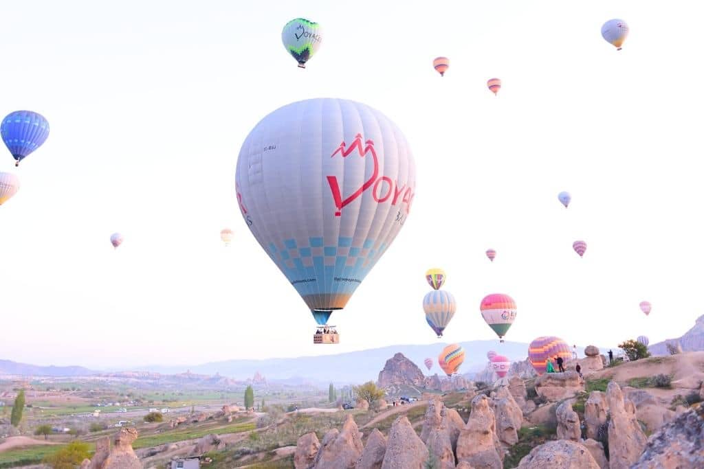 Colorful hot air balloons rise over the rocky terrain and fairy chimneys of Cappadocia during a serene morning. The largest balloon in the foreground bears the word 'Voyage' and is surrounded by smaller balloons drifting in the soft light. 