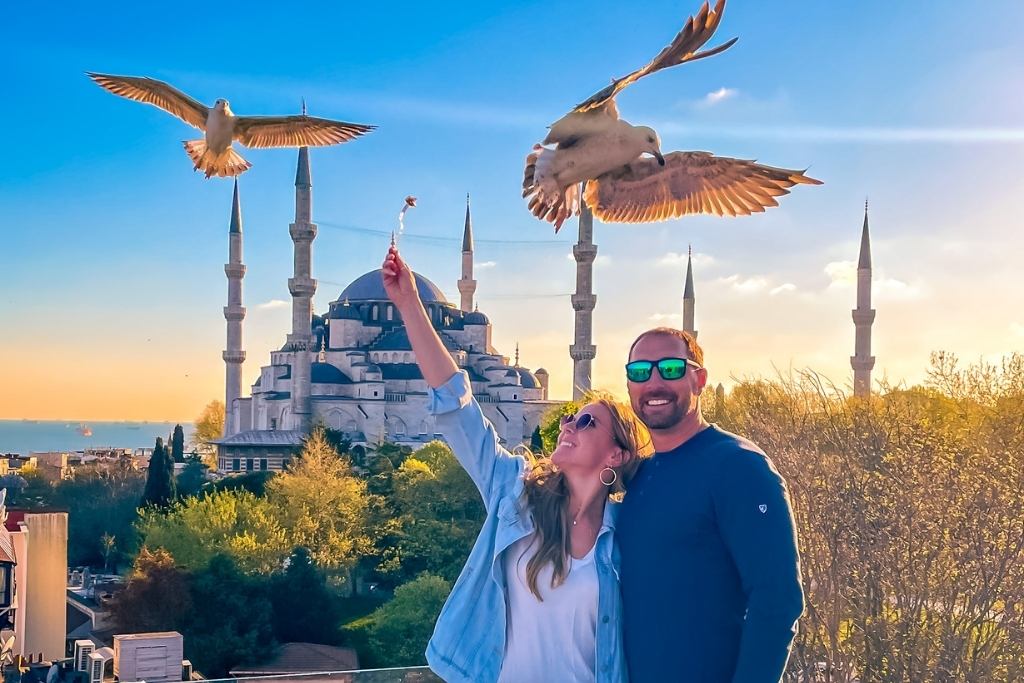 Kate and her husband pose joyfully in front of the Blue Mosque in Istanbul, with seagulls flying close overhead. The woman playfully holds up food to attract the birds, while the man smiles beside her. The scene is illuminated by warm sunlight, highlighting the mosque's iconic minarets and the surrounding greenery.