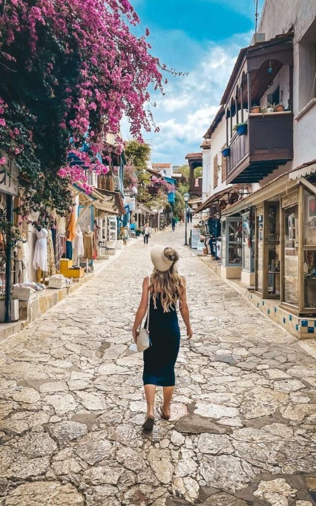 Kate in a black dress and wide-brimmed hat walks down a stone-paved street in Kaş, Turkey, lined with shops and vibrant bougainvillea draping from the buildings. The scene highlights the town's traditional Mediterranean charm."