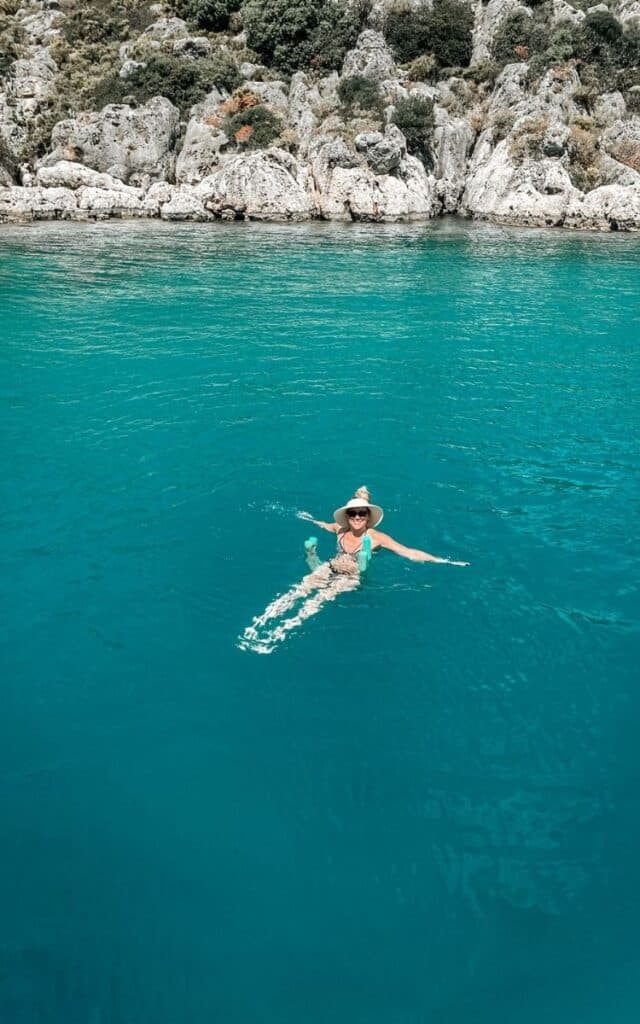 Kate swims in clear turquoise water near rocky cliffs on the Turkish Riviera in Turkey. She smiles at the camera as she floats on a pool noodle. 