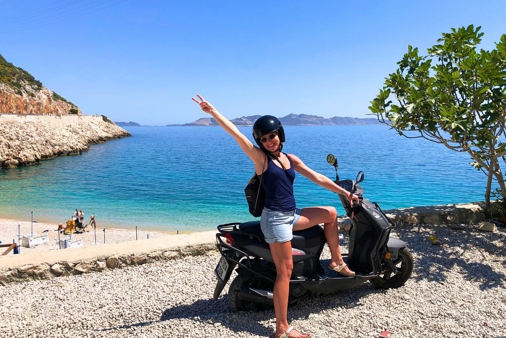 Kate poses on a scooter near a scenic beach on the Turkish Riviera, surrounded by turquoise waters, rocky cliffs, and a tree. She smiles and raises a peace sign under a bright blue sky.