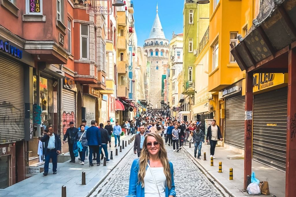 Kate stands on a bustling cobblestone street in Istanbul with the Galata Tower in the background, surrounded by colorful buildings and crowds of people. 